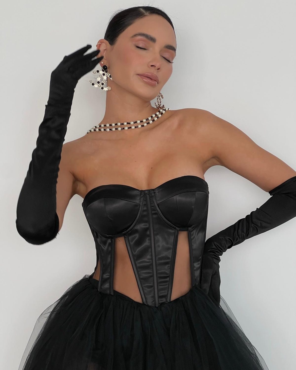 Jamillette Gaxiola in a black satin corset gown and long black gloves, with pearl jewellery, against a pale wall