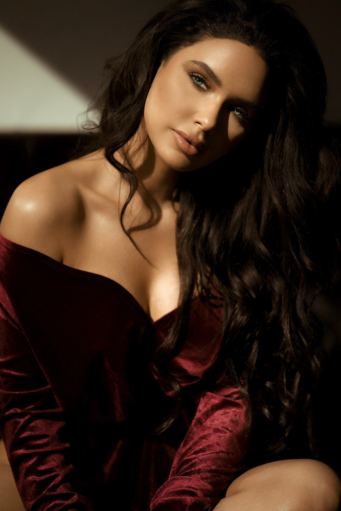 Portrait of Jamillette Gaxiola with long dark wavy hair, head tilted toward the camera, in an off-shoulder burgundy velvet top, lit by warm side light with deep shadow.