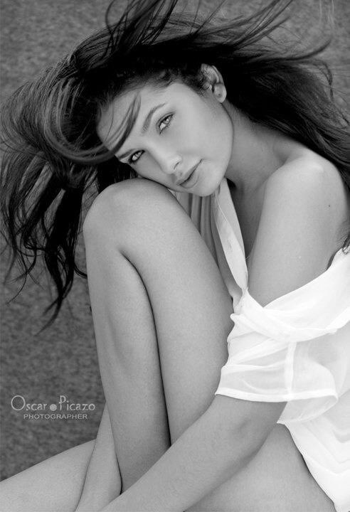 Black and white portrait of Jamillette Gaxiola seated with her cheek resting on her raised knee, dark hair flying, a loose white sheer blouse slipping off one shoulder. Signed Oscar Picazo, photographer.