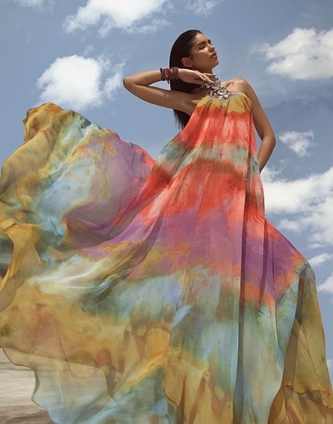 Jamillette Gaxiola photographed from below against a blue sky with white clouds, one arm raised with her hand at her jaw, in a bare-shouldered multicoloured gown billowing to one side.